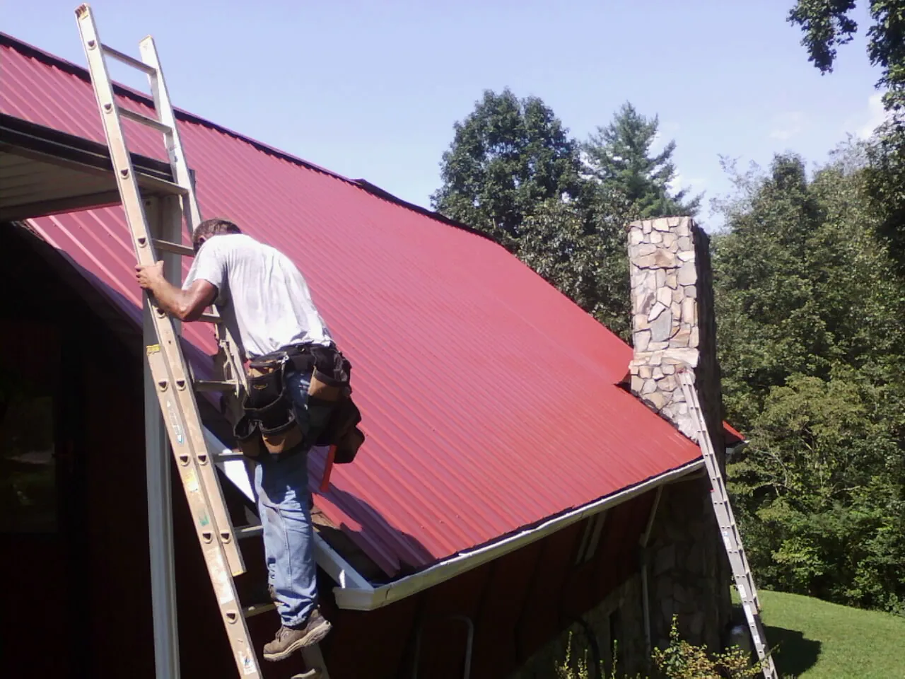 Roofer on a ladder beside a red metal roof and stone chimney — Soffit & Fascia Repair by Roofing Brothers in Eaton